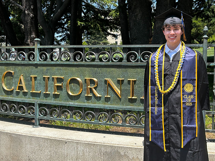 Image of Max in graduation regalia posing next to the UC Berkeley sign on campus